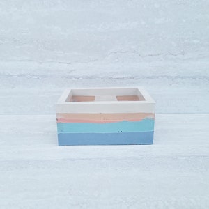 以下が含まれることがあります： 白の上面と多層デザインの正方形のマルチカラーコンテナ。層には、ライトピンク、ブルー、下部に濃いブルーが含まれます。コンテナはおそらくコンクリートまたは同様の材料で作られています。