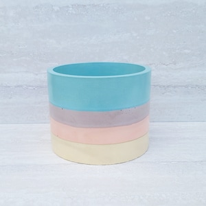 May include: A cylindrical, multi-colored planter with a pastel color scheme. The planter is composed of four horizontal bands: light blue, lavender, peach, and pale yellow. The planter is made of a solid material and has a smooth finish.