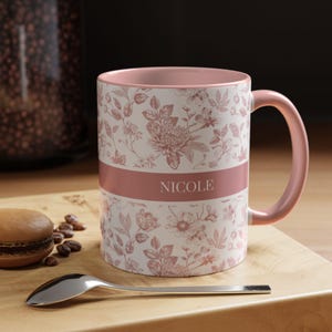 May include: A pink ceramic mug with a white floral pattern and a pink band with the name "NICOLE" printed on it.