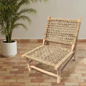 May include: A natural-toned woven chair with a light wood frame. The seat and back are made of a tightly woven rope material. The chair is set on a brick-patterned floor, with a potted plant in the background.
