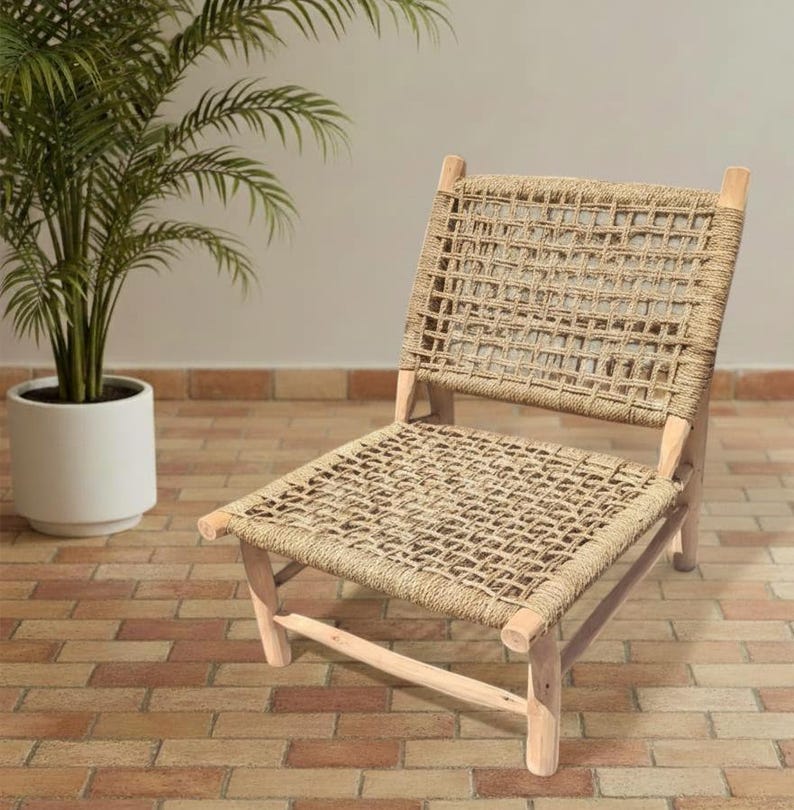 May include: A natural-toned woven chair with a light wood frame. The seat and back are made of a tightly woven rope material. The chair is set on a brick-patterned floor, with a potted plant in the background.