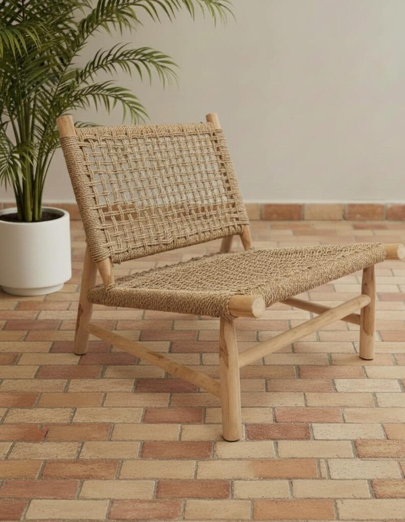 May include: A natural-toned woven rope chair with a wooden frame. The chair features a rectangular back and seat, with the rope woven in a grid pattern. The chair is set on a brick-paved surface, with a potted plant in the background.