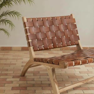 May include: A modern lounge chair with a woven leather seat and backrest. The chair features a light-colored wooden frame and a brown leather weave. The design is minimalist and suitable for indoor or outdoor use.