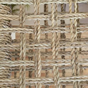 May include: Close-up of a woven natural fiber material with a grid-like pattern. The texture is a light beige color, with thick, braided strands creating a textured surface. The background shows a glimpse of a brick wall.