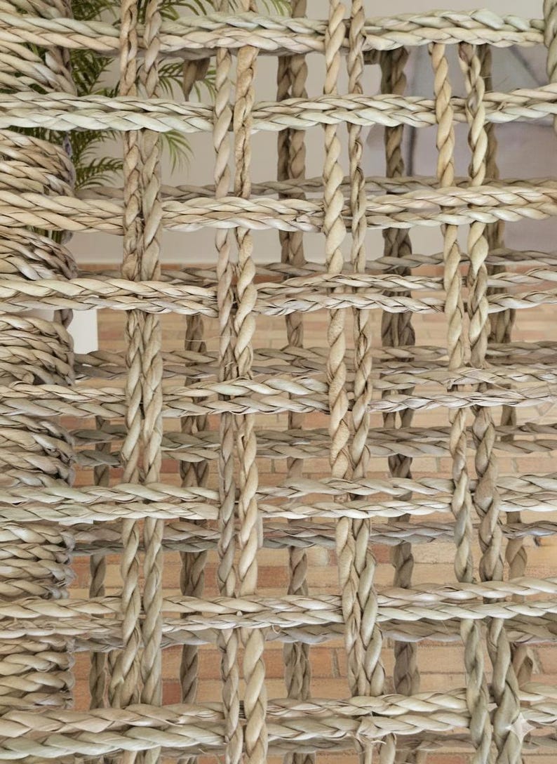 May include: Close-up of a woven natural fiber material with a grid-like pattern. The texture is a light beige color, with thick, braided strands creating a textured surface. The background shows a glimpse of a brick wall.