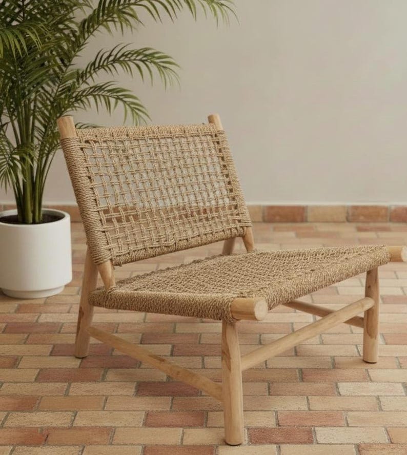 May include: A natural-toned woven rope chair with a wooden frame. The chair features a rectangular back and seat, with a woven rope design. The chair is made of light-colored wood and is placed on a brick patio. A potted plant is in the background.