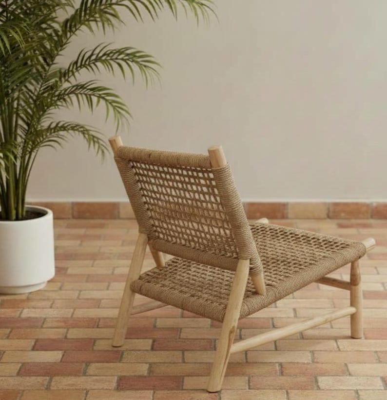 May include: A woven rope chair with a light wood frame. The chair has a woven seat and backrest, and is set against a neutral background. A potted plant is visible in the background.