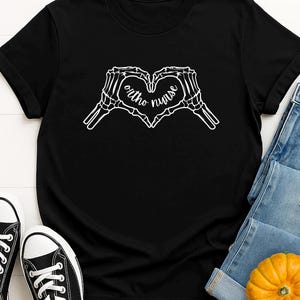 May include: Black t-shirt with a white skeletal hand heart design and the words "ortho nurse". The shirt is worn with blue jeans and black trainers. A small pumpkin is also visible.