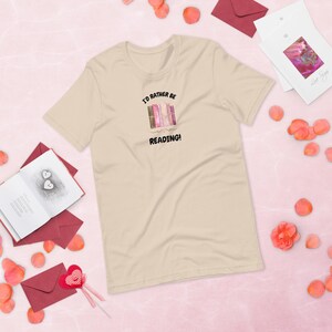 Peut inclure: T-shirt beige clair avec l'inscription « I'D RATHER BE READING! » au-dessus d'une illustration de livres. Autour du t-shirt, des enveloppes rouges, des pétales de rose et une carte de vœux.