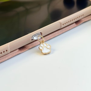 May include: A gold-colored phone charm with a white paw print design. The charm is attached to a clear plastic phone jack cover.