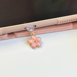 May include: A pink flower charm with a gold-colored bail is attached to a tablet case. The flower has five petals and a gold-colored outline. The tablet case is a light pink color.
