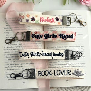 Peut inclure: Quatre porte-clés en tissu différents avec des motifs sur le thème du livre. Les porte-clés sont blancs avec du texte noir et présentent les phrases "Bookish", "Cute Girls Read", "Cute Girls Read Books" et "Book Lover".