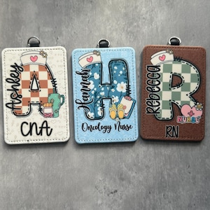 Peut inclure: Trois porte-badges personnalisés avec une boucle métallique en haut. Le premier est blanc avec un "A" à carreaux marron et blanc et les noms "Ashley" et "CNA". Le second est bleu clair avec un "H" floral et les noms "Hannah" et "Oncology Nurse". Le troisième est marron avec un "R" à carreaux vert et blanc et les noms "Rebecca" et "RN".