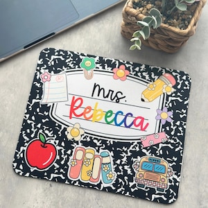 May include: A personalised mouse mat with a black and white composition notebook design. The mat features the name "Rebecca" in rainbow colours, surrounded by school-themed illustrations like an apple, pencil, and school bus.