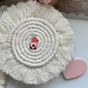 May include: A small, red and gold gnome charm with a white heart and white polka dots. The gnome is holding a red heart in its hands. The charm is on a white, round, crocheted coaster with fringe.