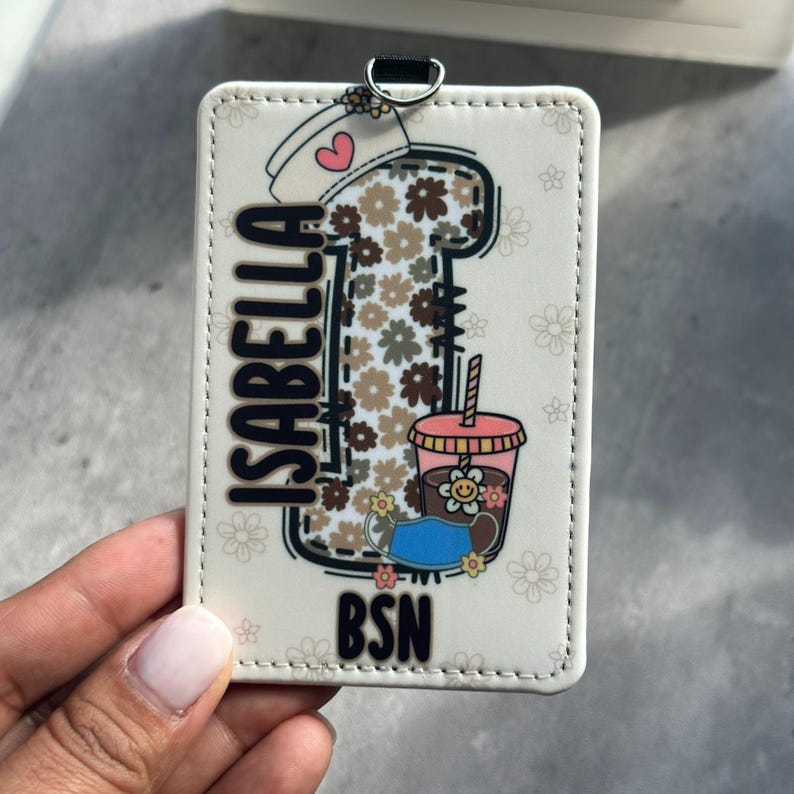 May include: A white badge holder with the name "ISABELLA" in black lettering. The design includes a coffee cup, a nurse's cap with a heart, and a drink with a flower. The letters "BSN" are at the bottom.