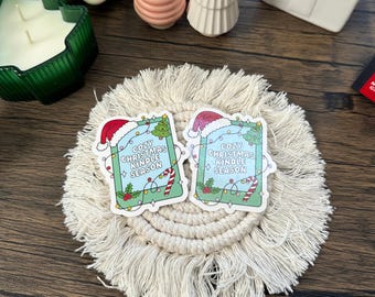 May include: Two stickers with the text "Cozy Christmas Kindle Season" featuring a Santa hat, Christmas tree, and candy cane design. The stickers are on a cream-colored macrame coaster, with other decorative items in the background.
