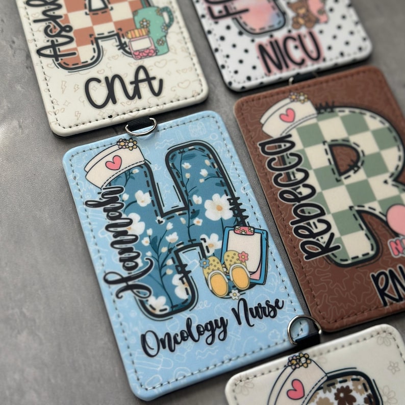 May include: A collection of personalized nurse badge reels. Each reel features a unique design, including floral patterns, initials, and the names of medical specialties like "Oncology Nurse" and "NICU". The reels are rectangular with a metal clip.