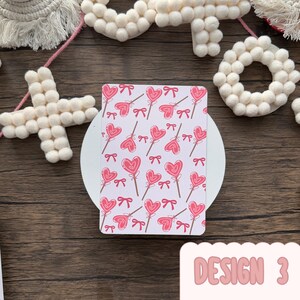 May include: A rectangular card featuring a pattern of pink heart-shaped lollipops and bows on a white backdrop. The text "DESIGN 3" is in pink, rounded font. The card is on a white circular surface. The background is a wooden surface.
