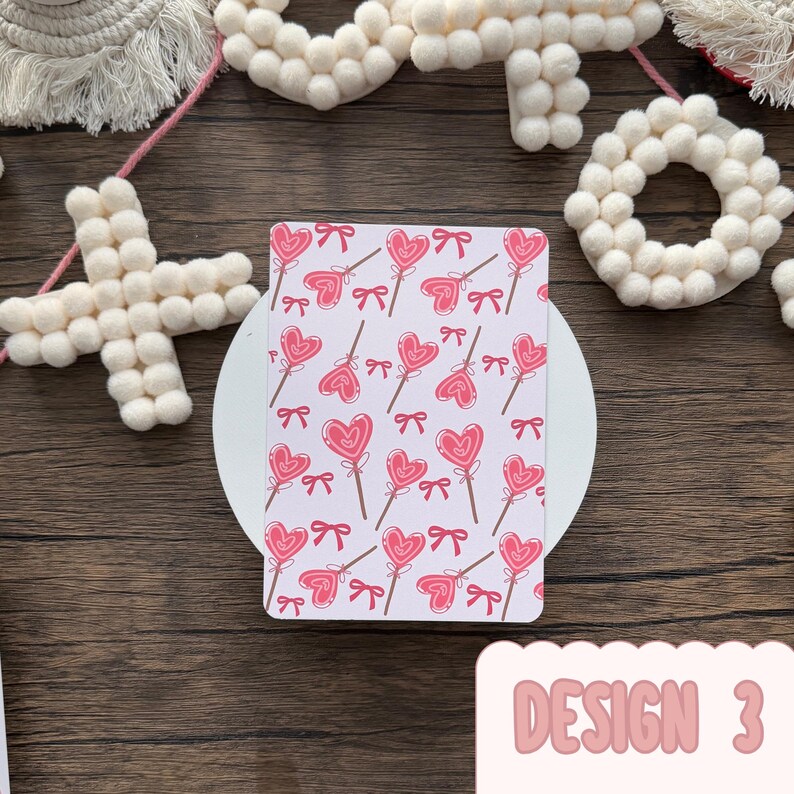 May include: A rectangular card featuring a pattern of pink heart-shaped lollipops and bows on a white backdrop. The text "DESIGN 3" is in pink, rounded font. The card is on a white circular surface. The background is a wooden surface.