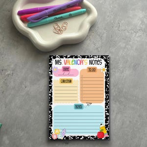 Pode incluir: Um bloco de notas colorido com o texto "MS. VALENCIA'S NOTES" e seções para data, lista de tarefas e notas. O bloco de notas tem uma borda preta e branca com estampa de vaca e é decorado com gráficos escolares. Canetas e clipes em uma bandeja branca.