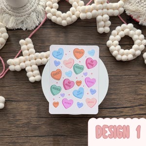 May include: A white rectangular card with candy heart illustrations in various colours, including orange, pink, and blue. The hearts have phrases such as "Sweet" and "Kisses." The card is labelled "Design 1." Decorative felt shapes are in the background.