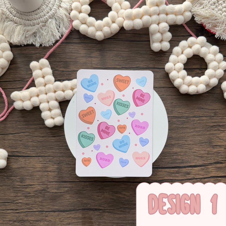 May include: A white rectangular card with candy heart illustrations in various colours, including orange, pink, and blue. The hearts have phrases such as "Sweet" and "Kisses." The card is labelled "Design 1." Decorative felt shapes are in the background.