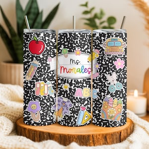 May include: Three stainless steel tumblers with a black and white composition notebook design. Each tumbler features colorful school-themed stickers, including an apple, school bus, and the name "Ms. Morales". The tumblers have silver straws.