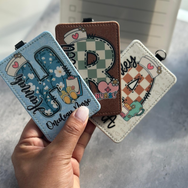 May include: Three personalized nurse badge holders with different designs. One is light blue with the name "Hannah" and the text "Oncology Nurse". Another is brown with a large letter "R" and the word "Nurse". The third is off-white with the name "Hey" and a triangle.