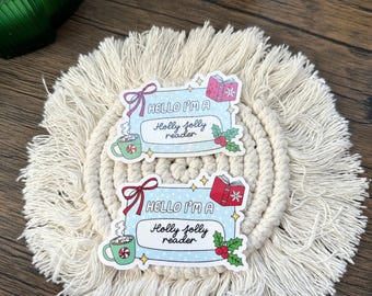 May include: Two stickers with the text "HELLO I'M A Holly jolly reader" are displayed on a cream-colored macrame coaster. Each sticker features a mug of hot cocoa, a book, and holly berries. The stickers have a light blue background.