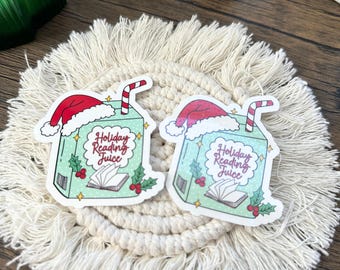 May include: Two stickers featuring a cartoon juice box design with a Santa hat, a striped straw, and the text "Holiday Reading Juice." The juice box is light blue with holly and berry accents. The stickers are on a cream-colored, macrame coaster.