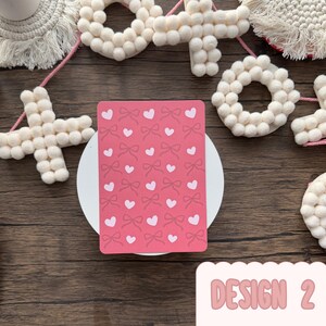 May include: A pink greeting card featuring a pattern of bows and hearts. The card rests on a white circular surface, surrounded by decorative white felt shapes and a wooden background. The text "DESIGN 2" is visible.