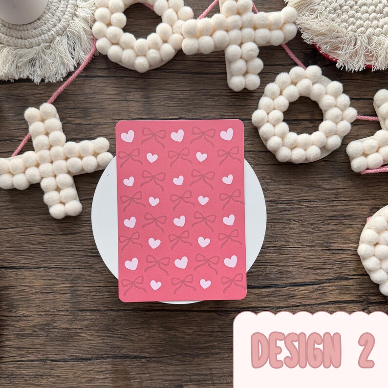 May include: A pink greeting card featuring a pattern of bows and hearts. The card rests on a white circular surface, surrounded by decorative white felt shapes and a wooden background. The text "DESIGN 2" is visible.