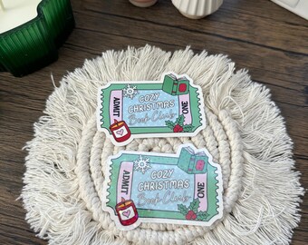 May include: Two stickers with a ticket design, featuring the text "COZY CHRISTMAS Book Club". The stickers have a light green border and include illustrations of a candle, a book, and holly. The background is a cream-colored, woven coaster.