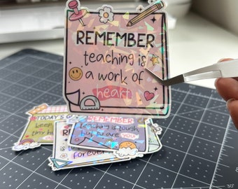 May include: A holographic sticker with the text "REMEMBER teaching is a work of heart" in pink and white lettering. The sticker has a rainbow iridescent background with a variety of small illustrations including a heart, a star, a pencil, a ruler, and a smiley face.