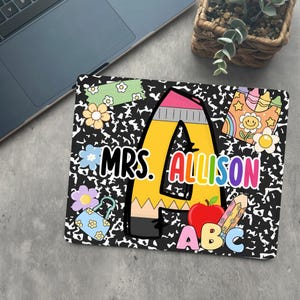 May include: A laptop sleeve with a black and white pattern, featuring a large yellow pencil-shaped letter "A" with the name "MRS. ALLISON". The design includes a red apple, a pencil, and floral and school-themed graphics.