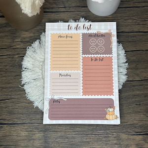 May include: A rectangular to-do list notepad with a gingham pattern background. The notepad features sections for main focus, mood tracker, priorities, to-do list, and notes. Decorative elements include ribbons and a pumpkin illustration. The text "to do list" is at the top.