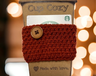 May include: A brown cardboard cup cozy with a red knitted sleeve and a wooden button. The cozy has the text "Cup Cozy" and "Made with love." printed on it.