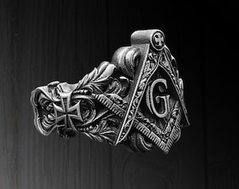 May include: A silver ring with Masonic symbols, including a square and compass with the letter "G". The ring has detailed engravings and a cross design. The ring is a piece of jewellery.