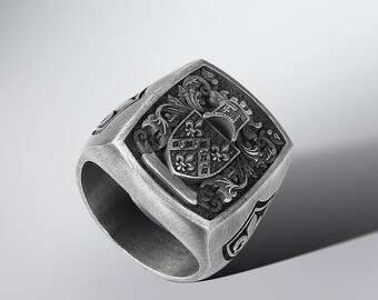 May include: A silver-toned ring with a square face featuring an ornate crest design. The ring has a thick band and detailed scrollwork around the crest. The ring is likely made of metal and is displayed on a white surface.