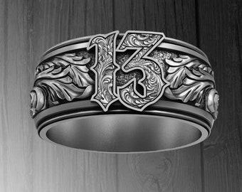Può includere: Un anello in metallo color argento con il numero 13 in un carattere decorativo e ornato. La fascia è dettagliata con intricati motivi a volute in rilievo simili a foglie che avvolgono i lati, su uno sfondo scuro e incavato.