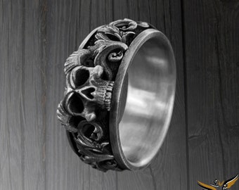May include: A silver ring with a detailed skull design. The ring is set against a dark wood background.