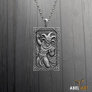 Puede incluir: Un colgante rectangular de plata en una cadena a juego que presenta un relieve detallado de un bufón con un sombrero de varias puntas y un cuello con volantes. El colgante está enmarcado por un borde trenzado intrincado, acabado oxidado.