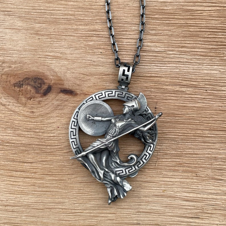 May include: A silver-toned pendant necklace featuring a detailed depiction of Athena, the Greek goddess of wisdom and warfare. The pendant showcases Athena with a shield and spear, encircled by a decorative border, suspended from a matching chain.