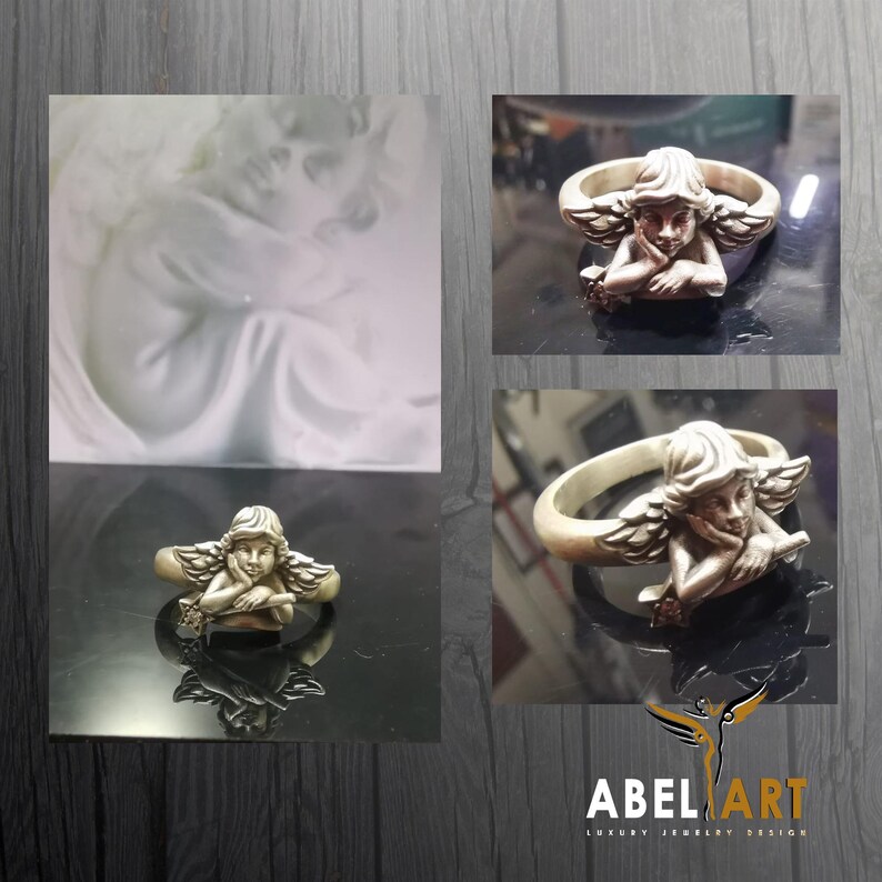 May include: A silver-toned ring featuring a detailed angel design. The angel is depicted with wings and is resting its head on its hand. The ring has a smooth band and a detailed angel figure. The ring is displayed on a reflective surface.