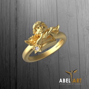 May include: A gold-colored ring featuring a sculpted angel resting on the band. The angel has detailed hair and wings, with a star-shaped accent holding a small, clear gemstone. The ring is set against a dark wood-grain background.