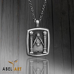 May include: Silver pendant necklace with a square-shaped charm featuring a Masonic symbol with two pillars, a compass and square, and an all-seeing eye.
