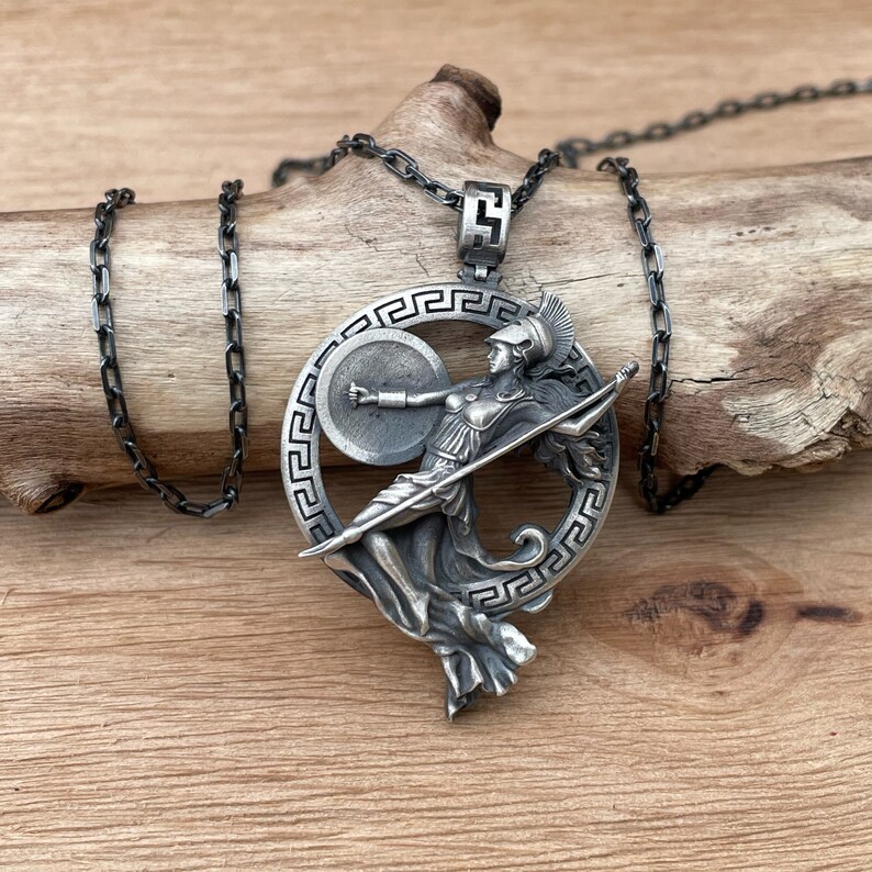 May include: A silver-toned pendant necklace featuring a detailed depiction of Athena, the Greek goddess of wisdom and warfare. The pendant is set within a circular frame with a Greek key design, and hangs from a dark chain.