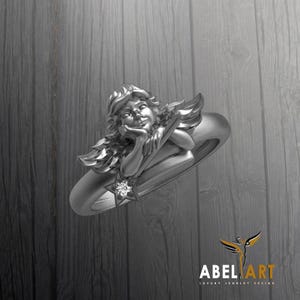 May include: A silver ring featuring a detailed angel design. The angel is depicted with flowing hair, wings, and a star-shaped accent with a central stone. The ring has a polished finish and a classic band.