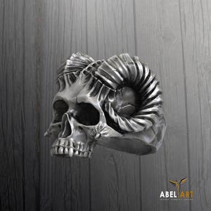 Può includere: Un anello in argento a forma di teschio con corna di ariete. L'anello è decorato con sculture intricate e una finitura testurizzata.